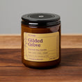Load image into Gallery viewer, An amber glass jar candle with a black lid and a tan label reading 'Gilded Grove' by Elm Design, placed on a wooden surface against a neutral gray background.
