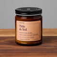 Load image into Gallery viewer, Tide & Sol scented soy candle in amber glass jar on wooden surface.
