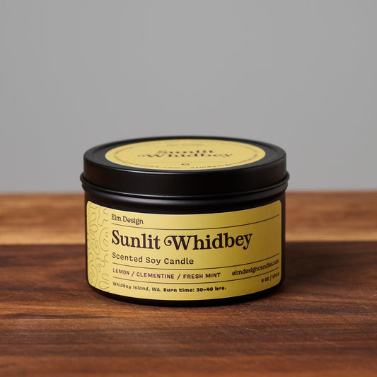 Sunlit Whidbey scented soy candle in black metal tin on wooden surface.