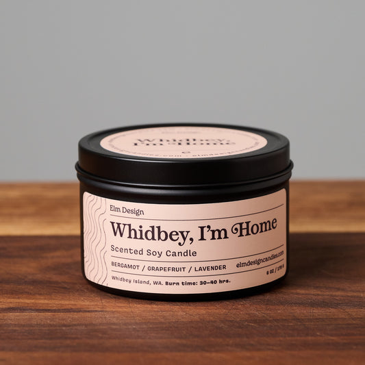 Whidbey, I'm Home scented soy candle in black metal tin on wooden surface.