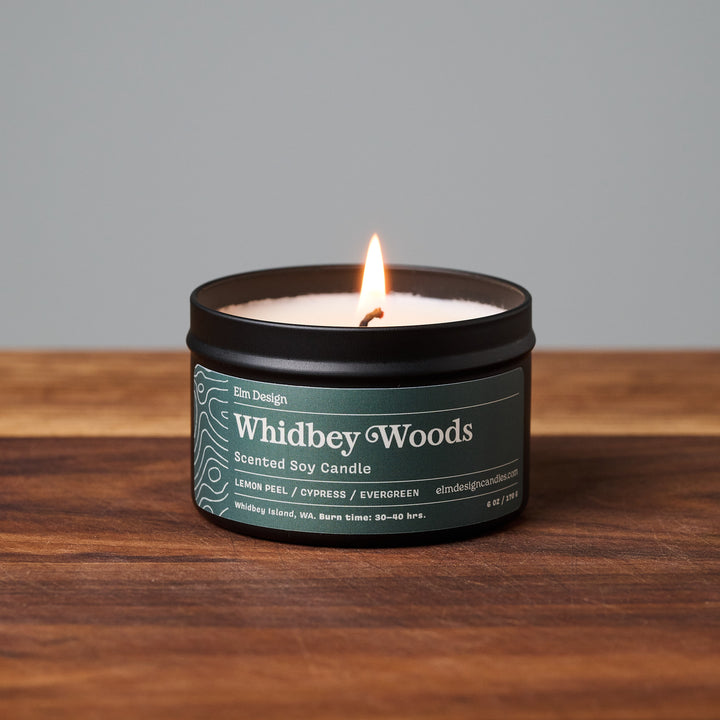 Whidbey Woods scented soy candle in black metal tin on wooden surface.