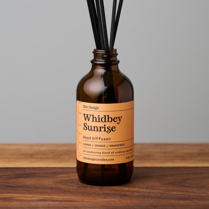 Whidbey Sunrise scented reed diffuser in amber glass jar with black reeds on wooden surface.
