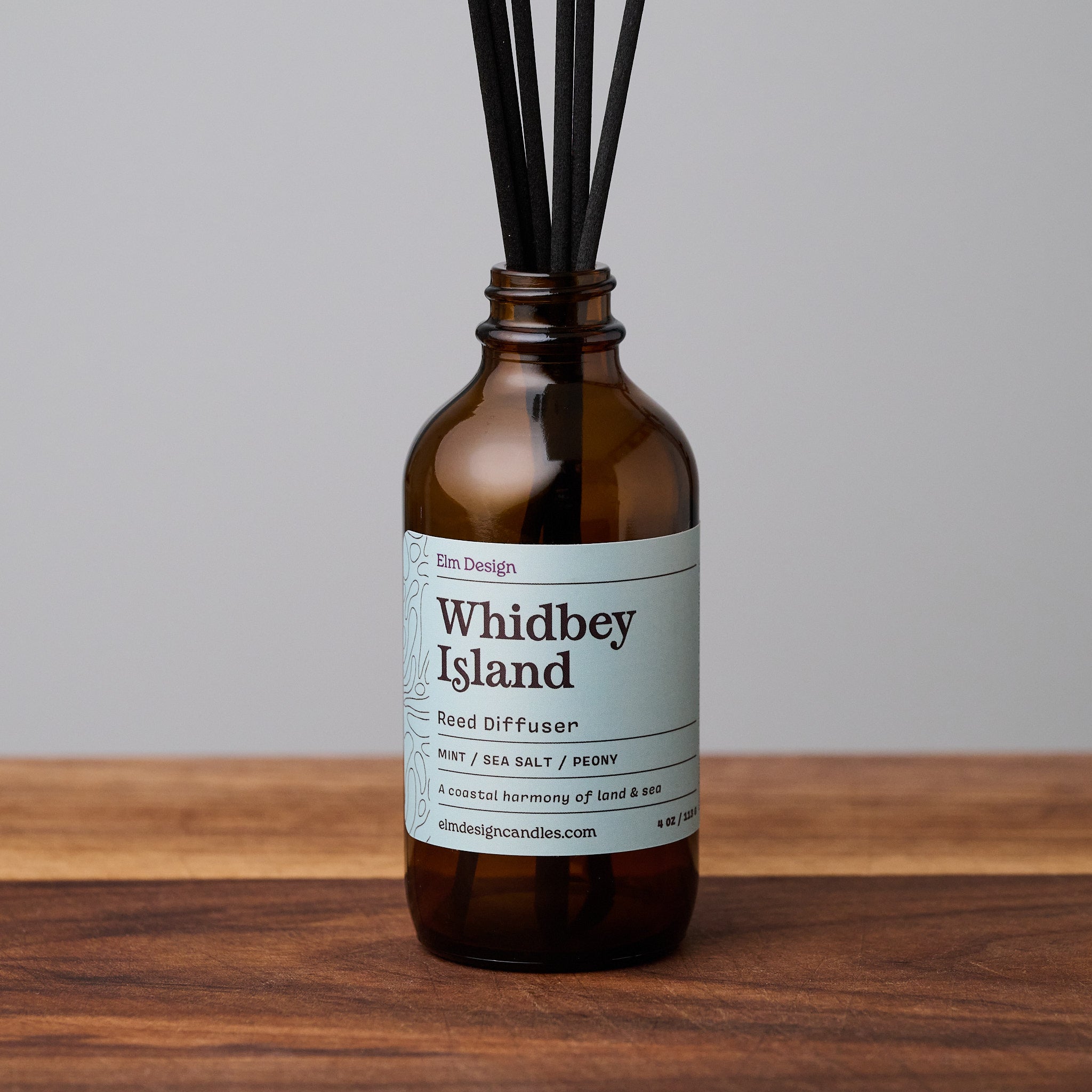 Whidbey Island scented reed diffuser in amber glass jar with black reeds on wooden surface.