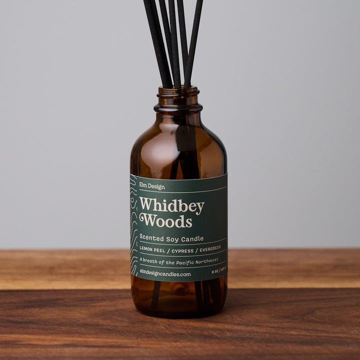 Whidbey Woods scented reed diffuser in amber glass jar with black reeds on wooden surface.