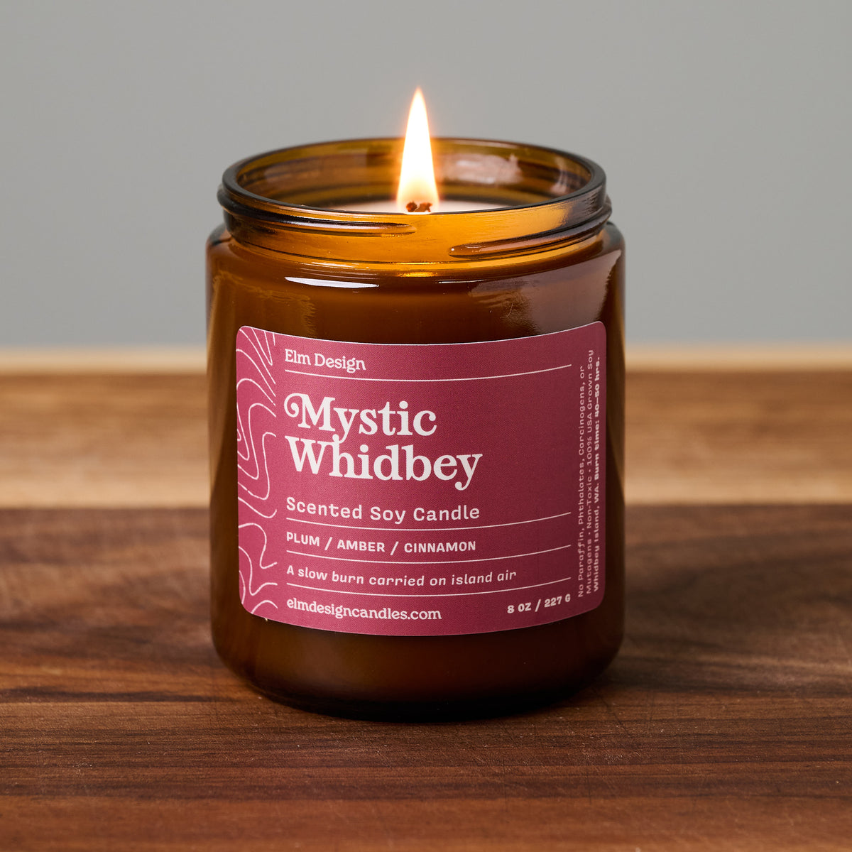 An amber glass jar candle with a lit wick, featuring a plum-colored label that reads 'Mystic Whidbey' by Elm Design, set on a wooden surface against a neutral gray background.