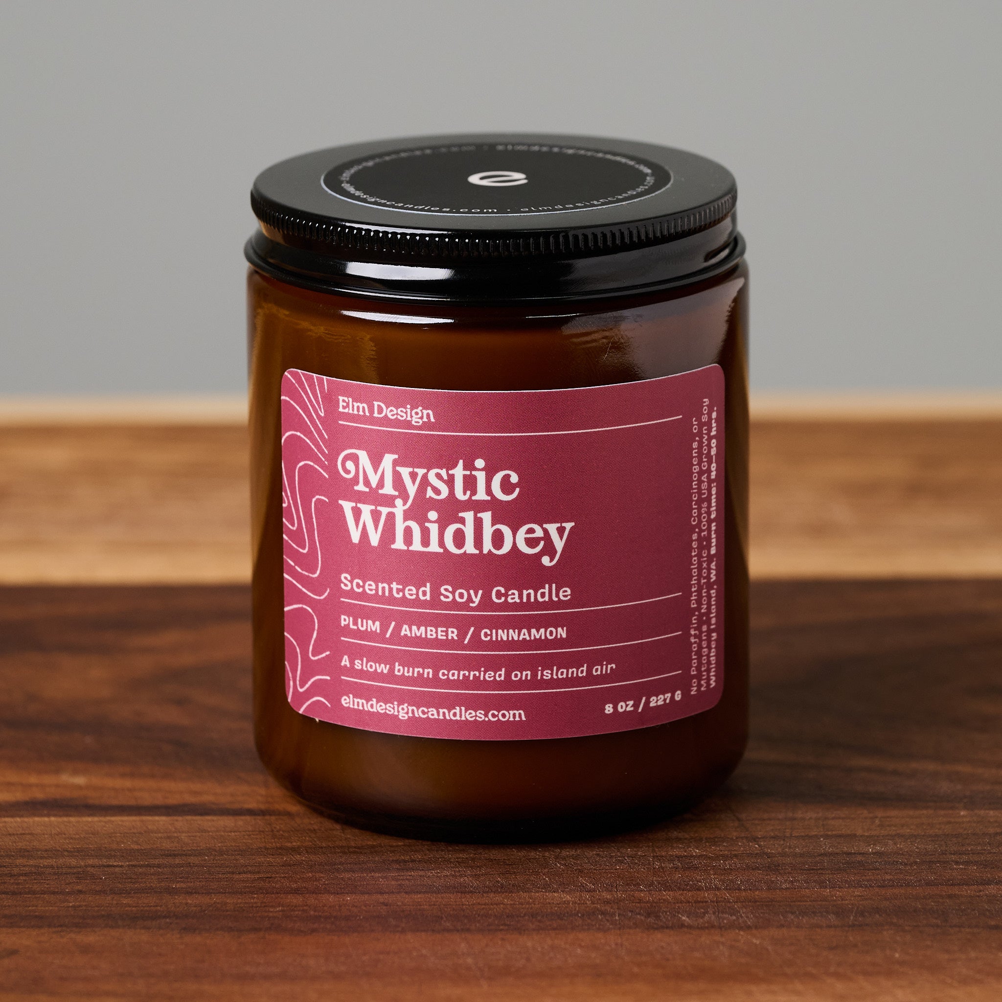 An amber glass jar candle with a black lid and a dusty rose label reading 'Mystic Whidbey' by Elm Design, placed on a wooden surface against a neutral gray background.