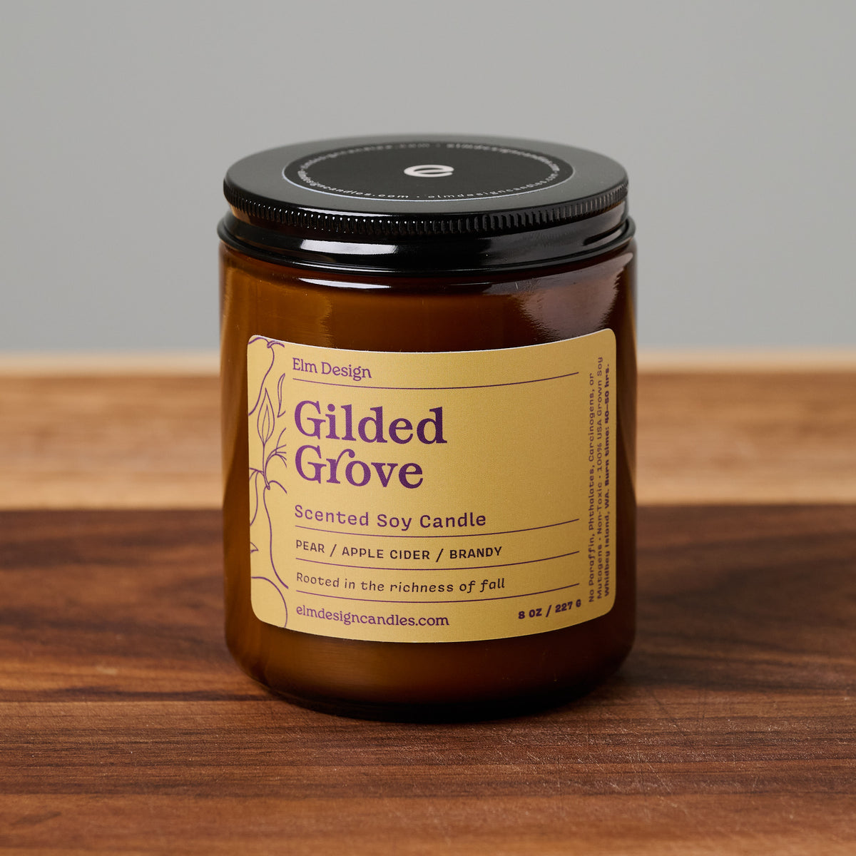 An amber glass jar candle with a black lid and a tan label reading 'Gilded Grove' by Elm Design, placed on a wooden surface against a neutral gray background.