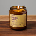 Load image into Gallery viewer, A lit amber glass jar candle labeled 'Gilded Grove' by Em Design, placed on a wooden surface against a neutral gray background.
