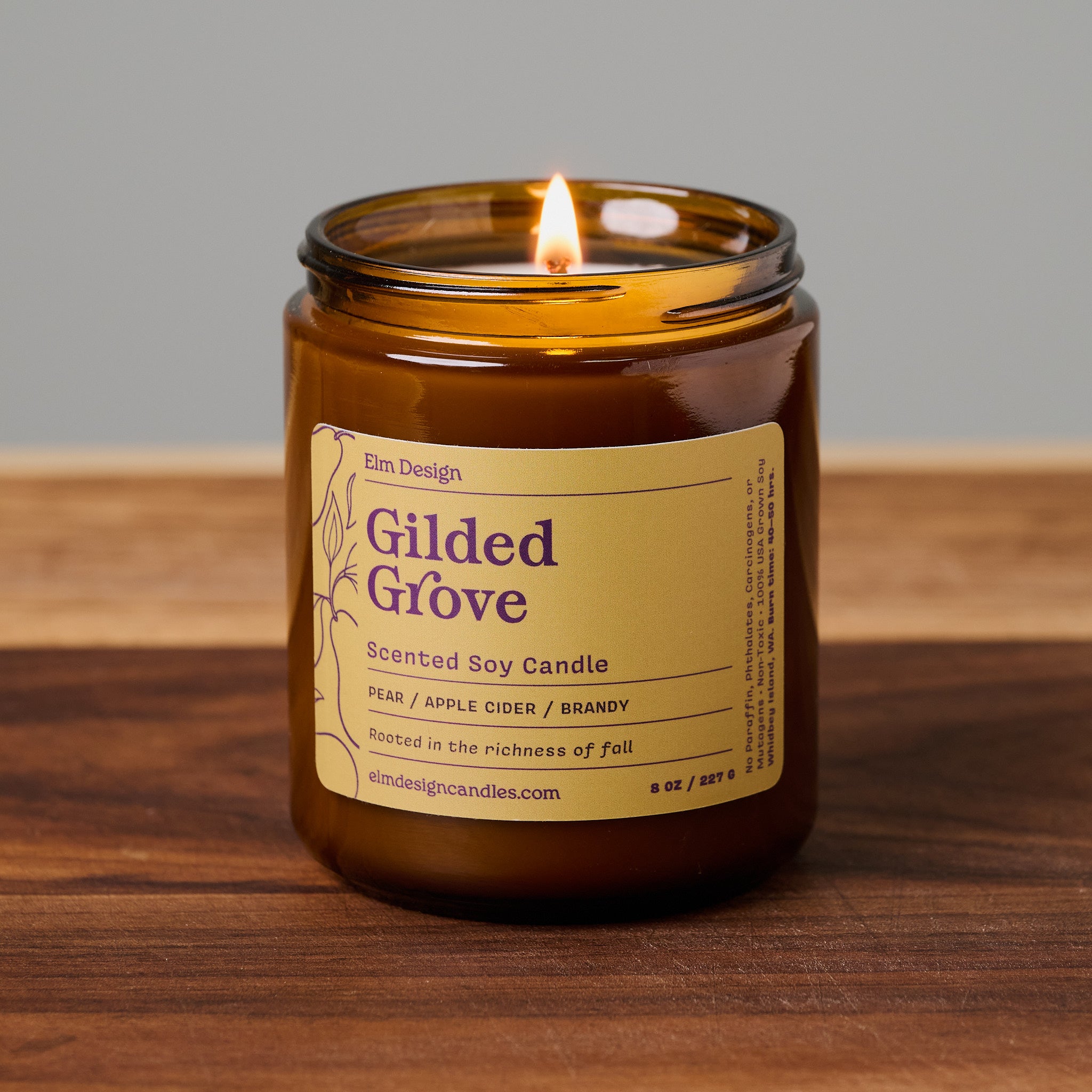 A lit amber glass jar candle labeled 'Gilded Grove' by Em Design, placed on a wooden surface against a neutral gray background.