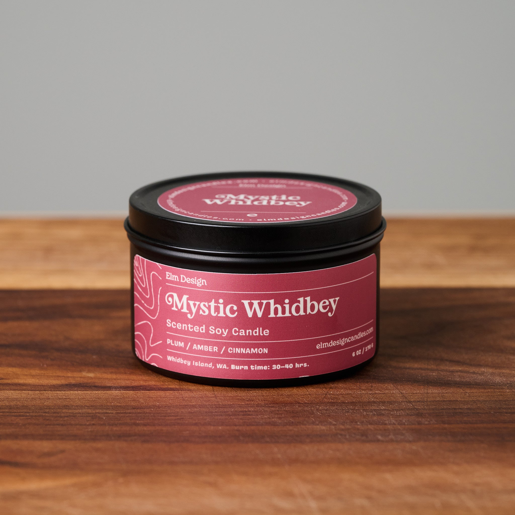 A black metal tin candle with a dark pink label reading 'Mystic Whidbey' sits on a wooden surface against a light gray background.
