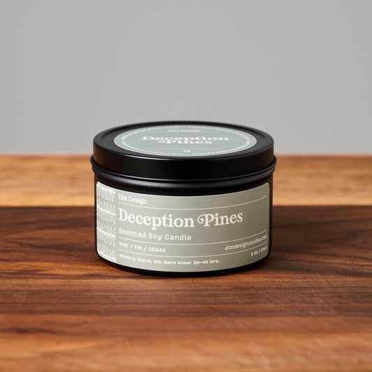 A black metal tin candle labeled 'Deception Pines' sits on a wooden surface against a neutral gray background.