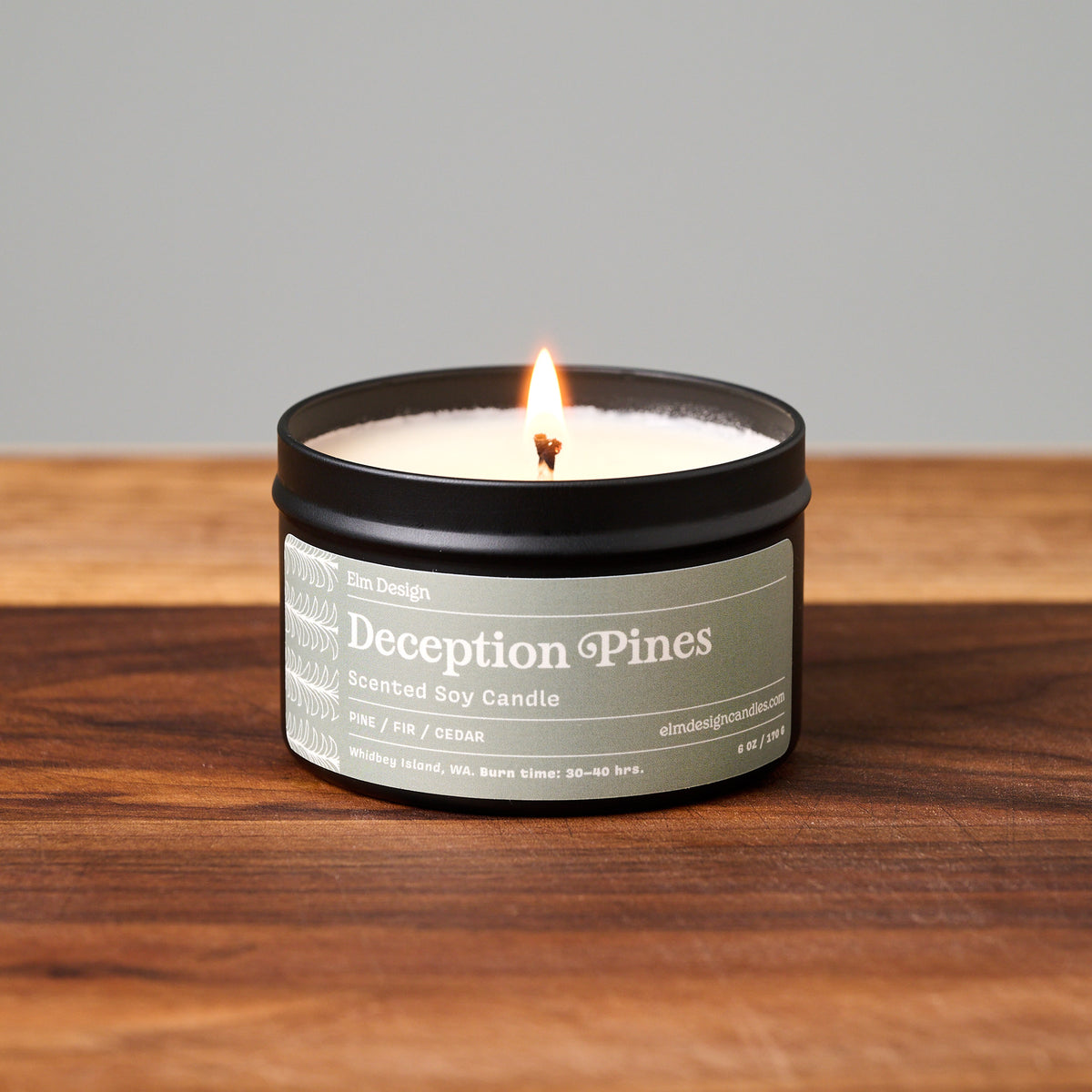 A lit black metal candle tin on a wooden surface against a neutral gray background. The label reads 'Deception Pines' and 'scented soy candle'.