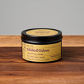 Load image into Gallery viewer, Gilded Grove scented soy candle in a black metal tin with a gold label, resting on a wooden surface against a neutral gray background.
