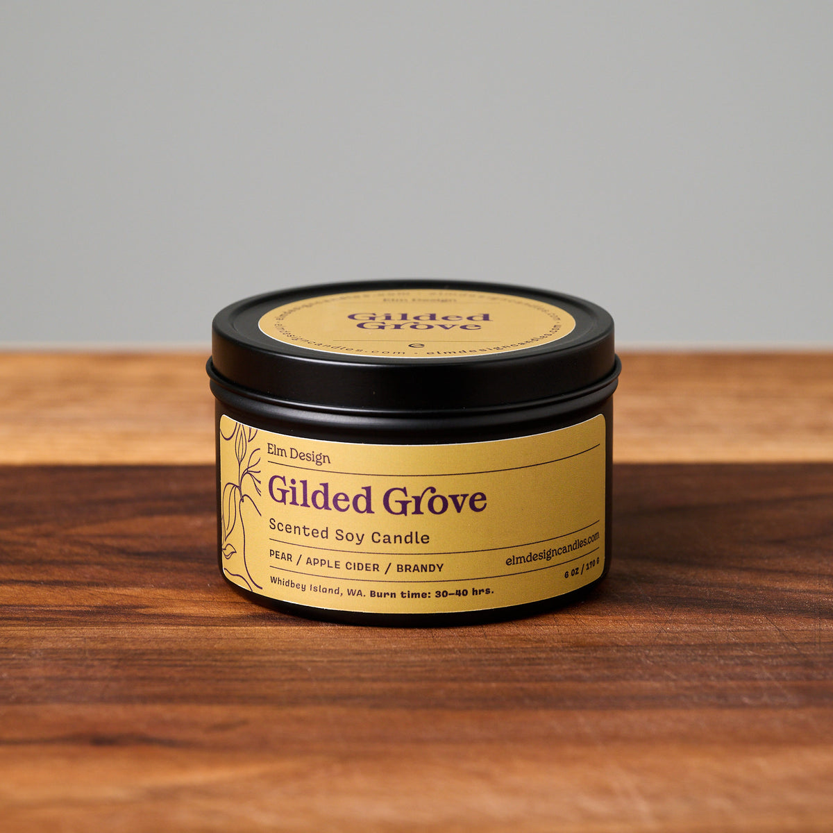 Gilded Grove scented soy candle in a black metal tin with a gold label, resting on a wooden surface against a neutral gray background.