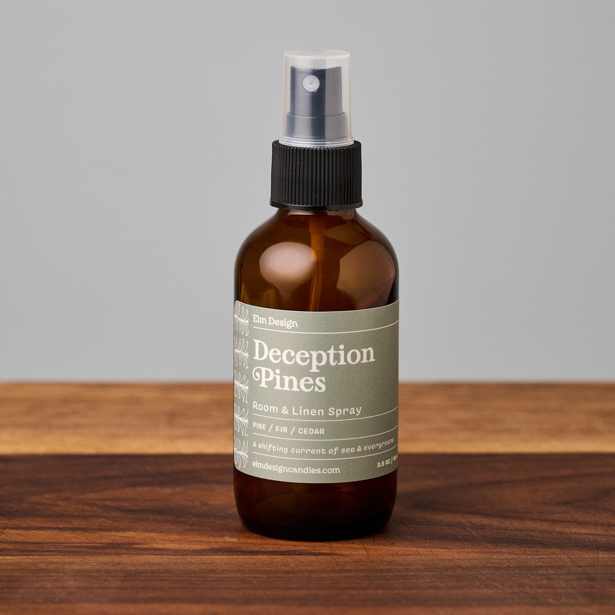 Scented room spray in Deception Pines scent. Amber glass bottle with a black lid sitting on a wooden cutting board with a light gray background.