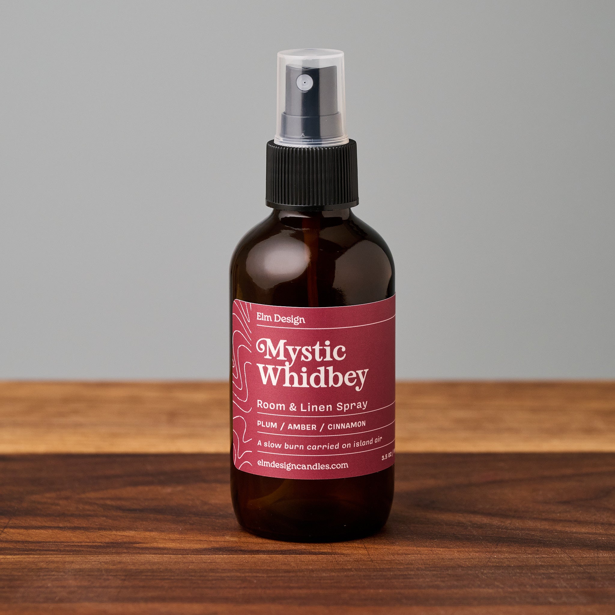An amber glass room and linen spray bottle with a black spray top and a dark pink label reading 'Mystic Whidbey' by Elm Design, standing on a wooden surface against a plain gray background.