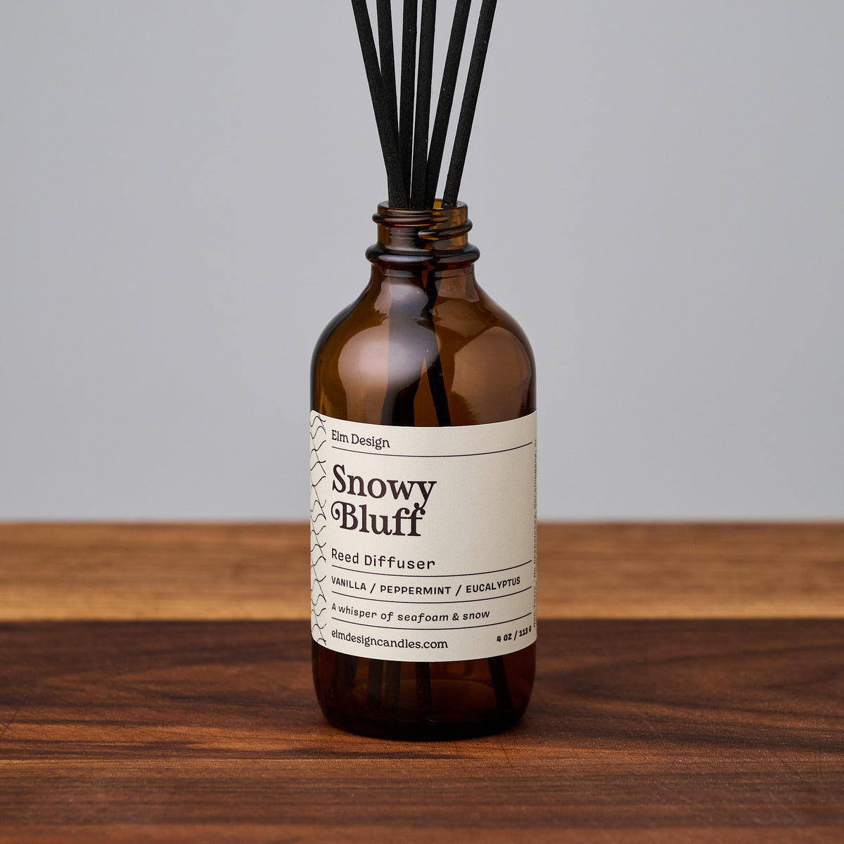 An amber glass reed diffuser bottle with black reeds and a cream label reading 'Snowy Bluff' by Elm Design, placed on a wooden surface against a light gray background.