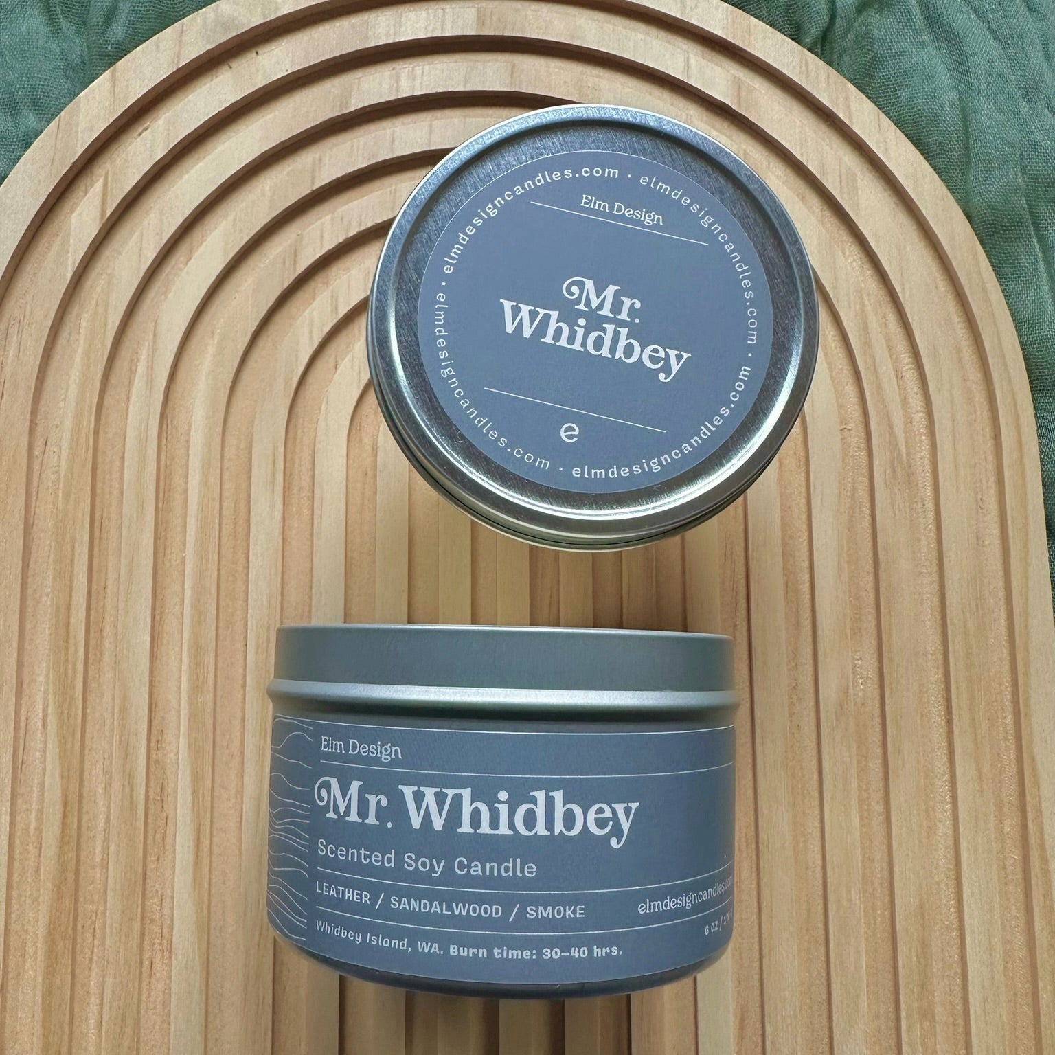 Two 'Mr. Whidbey' candles on a wooden surface with a green fabric background