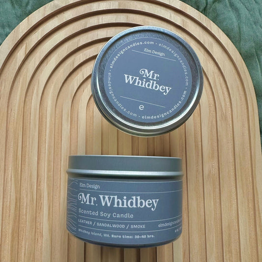 Two 'Mr. Whidbey' candles on a wooden surface with a green fabric background
