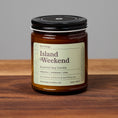 Load image into Gallery viewer, Island Weekend scented soy candle in amber glass jar on wooden surface.
