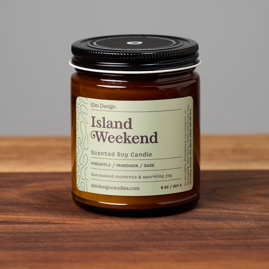 Island Weekend scented soy candle in amber glass jar on wooden surface.