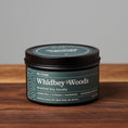 Load image into Gallery viewer, Whidbey Woods scented soy candle in black metal tin on wooden surface.
