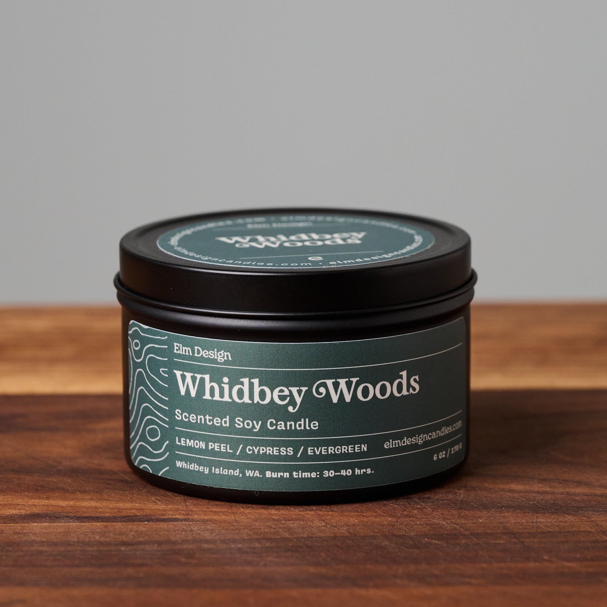 Whidbey Woods scented soy candle in black metal tin on wooden surface.