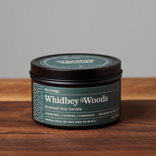 Whidbey Woods scented soy candle in black metal tin on wooden surface.